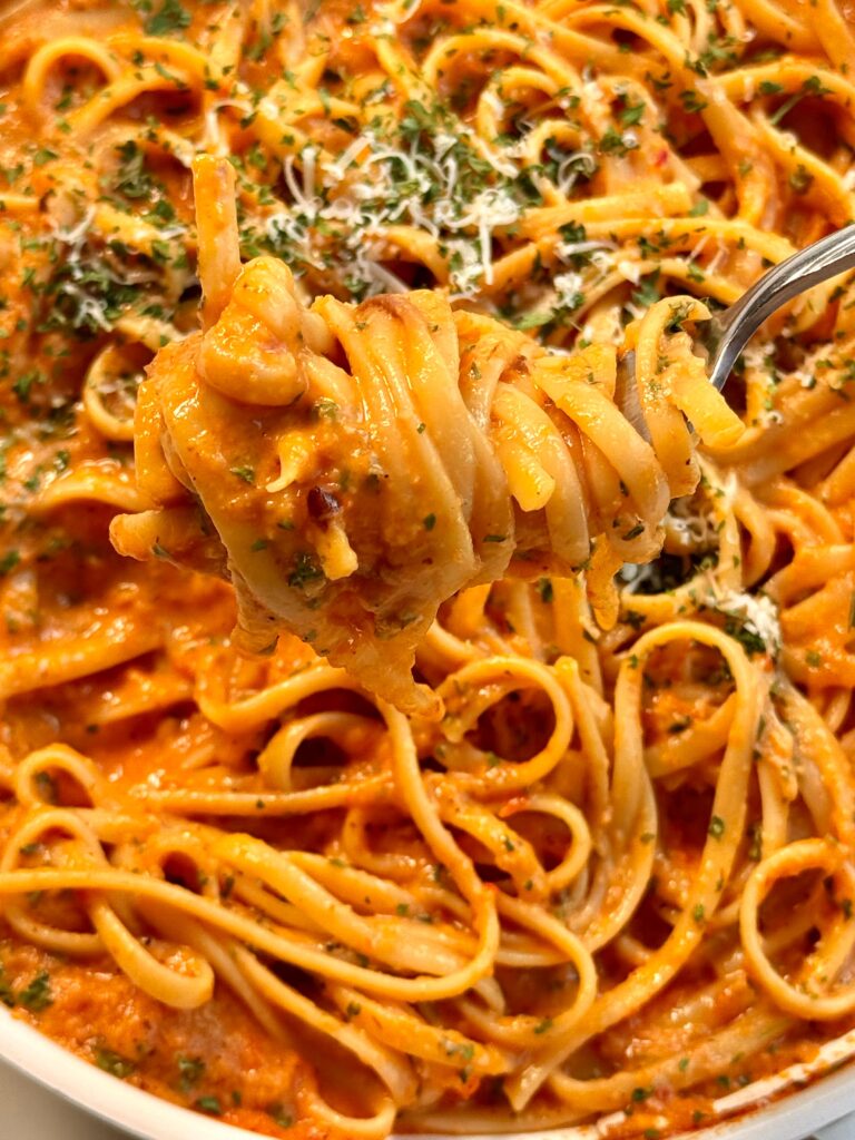 Close-up of a fork twirling creamy red pepper pasta, showing linguine coated in a rich roasted red bell pepper sauce, topped with freshly grated parmesan cheese and a sprinkle of chopped parsley for garnish.