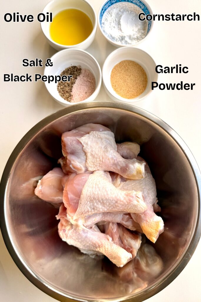 Raw chicken drumsticks in a metal bowl surrounded by small bowls of olive oil, cornstarch, garlic powder, and a mix of salt and black pepper, ready for seasoning.