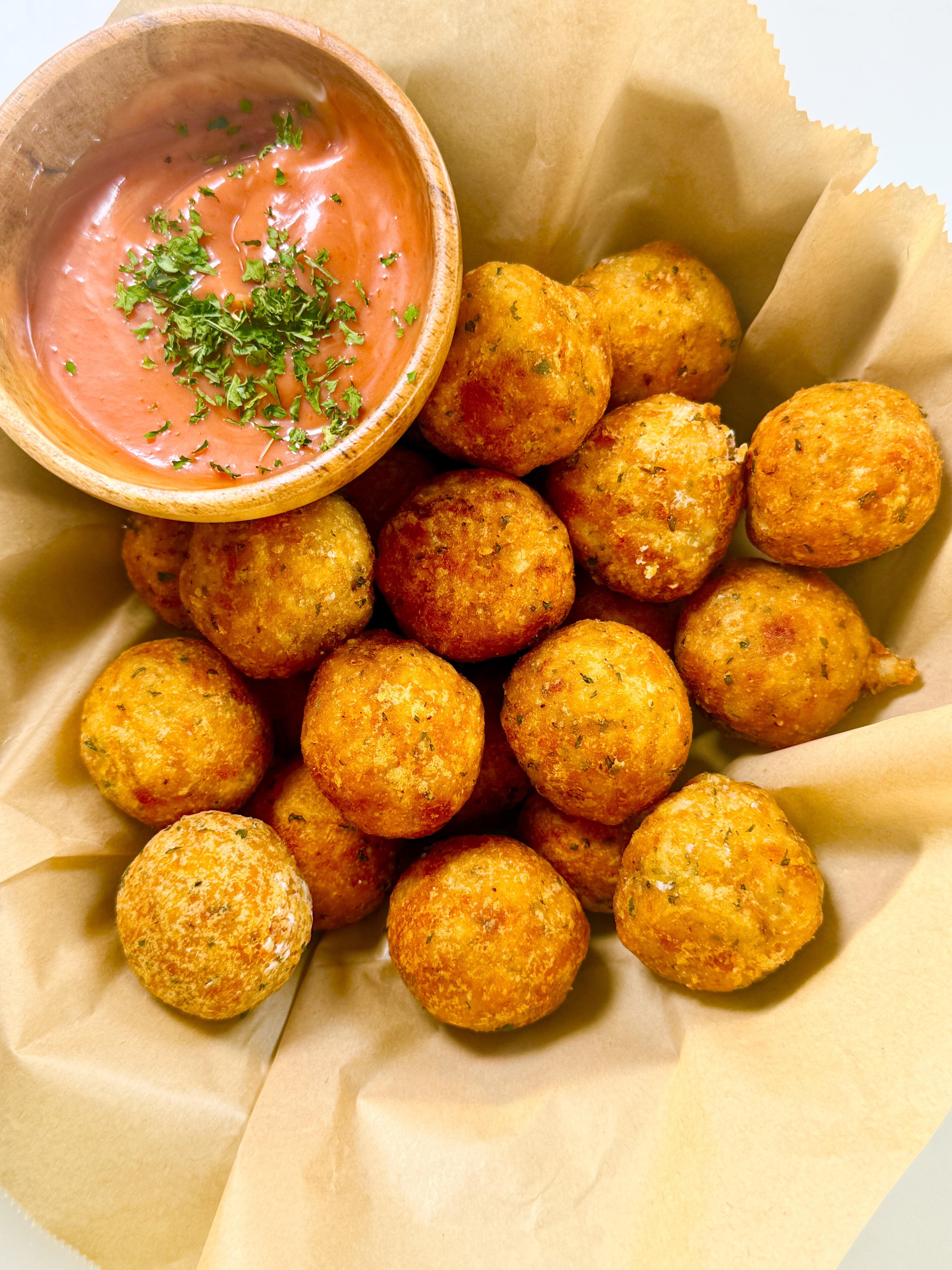 Overhead shot of fried potato balls in parchment paper with a wooden bowl of ketchup mayo dip garnished with parsley