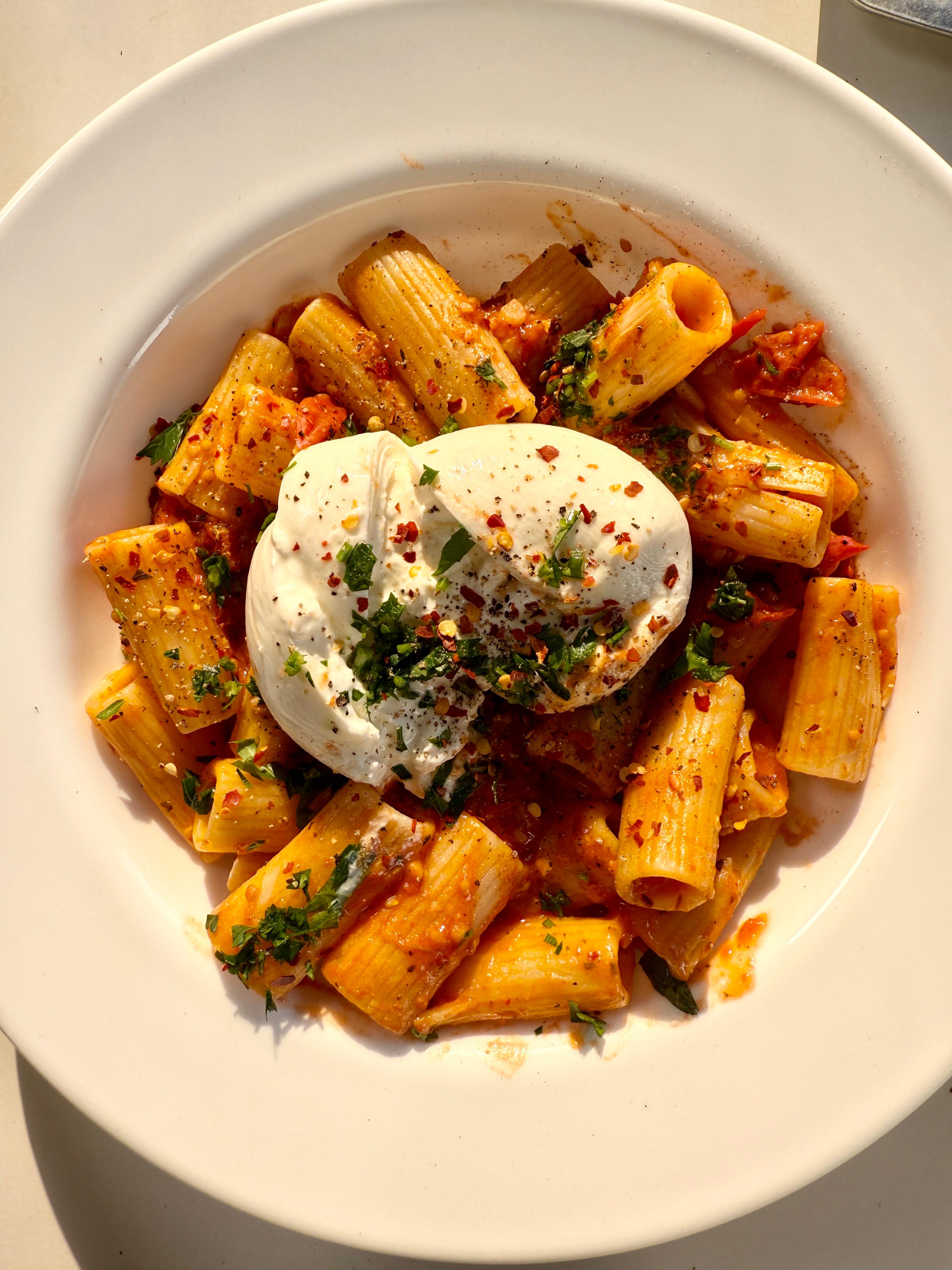 Overhead view of creamy burrata sitting on saucy rigatoni pasta, sprinkled with parsley and red pepper flakes