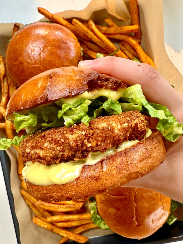 Hand holding a crispy homemade McChicken sandwich with lettuce and honey mustard mayo, served with seasoned fries in the background